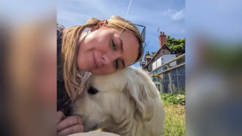 Rhianne Aitken smiling with a golden retriever.  A fence and a house are in the background. The image was taken on a sunny day.
