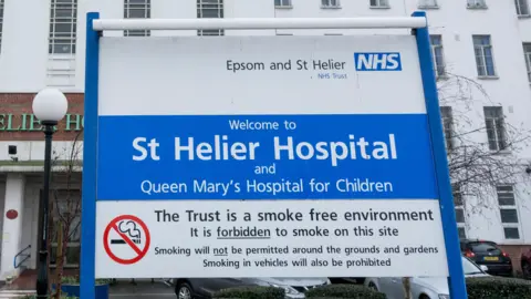 Exterior of St Helier Hospital shows white building entrance with blue and white Epsom and St Helier NHS trust sign welcoming patients and stating it is smoke free