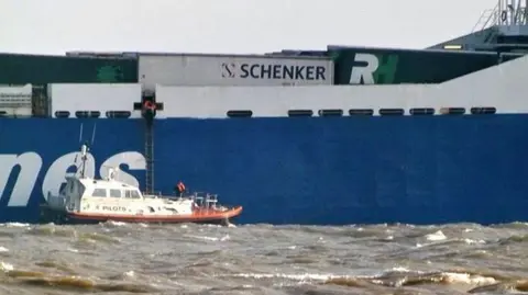 A small boat is situated in what looks like choppy sea next to a port with a ladder suspended from it which goes up into a larger platform.