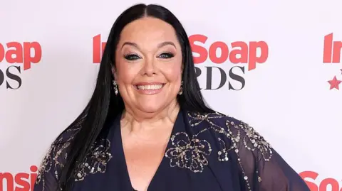 Getty Images A photo of Lisa Riley from the shoulders up. She has long dark hair and is wearing a blue dress with beaded embellishments, and is standing in front of a background with the logo for the Soap Awards. She is smiling at the camera.