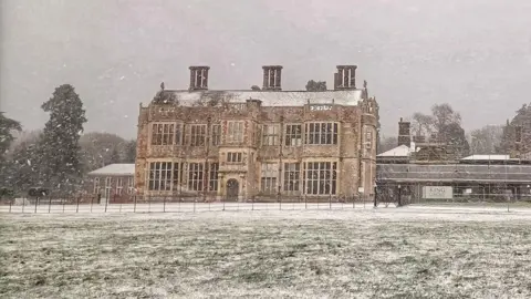 Norfolkwalkabout/WeatherWatchers Felbrigg Hall is an old two-storey building with large chimneys. There is a grass in front of the building, which has snow starting to settle on it.