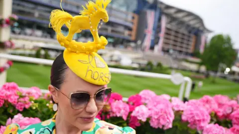 A woman wearing a yellow fascinator which has a model of a rocking horse at the top of it and "I [love] horses" studded into it.