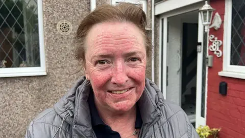 Head and shoulders of Gill Thornton standing outside her home. She is smiling and has short brown hair and is wearing a grey padded coat