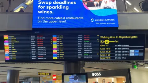 Two departure boards at Gatwick airport on 28 December, showing status of flights, some of which are delayed. A third board shows the walking time to reach various departure gates. In the background, there are two stores - a Lego store and a Hugo Boss store.