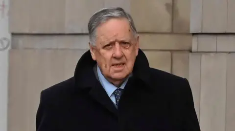 William Lloyd-Lavery, a man with grey hair, a dark jacket, blue shirt and blue tie, pictured beside a grey brick building. He has his mouth slightly ajar.