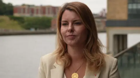 Kim McGuinness, who has long blonde hair, stands on the banks of the River Tyne, she is dressed in a beige suit with a gold medallion necklace hanging from her neck.