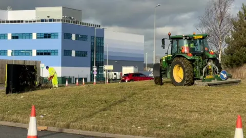 Martin Heath/BBC A grass-covered roundabout on a main road, with a green tractor pulling a weeding device. A man in yellow PPE can be seen to the left of the picture with a spraying tool. There are road cones around the edge of the roundabout and a three-storey blue office building to the left.