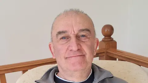 David Harris David Harris has short grey hair and is looking at the camera. He is clean shaven. There is a staircase banister behind him. He is wearing a grey fleece and a black top. 