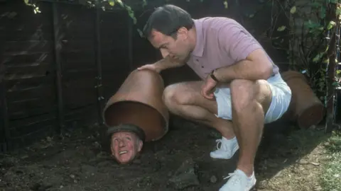 Actor Angus Dayton lifting a flowerpot to reveal Richard Wilson buried up to his neck in a back garden in an episode of One Foot in the Grave