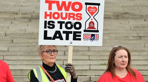 Two women side by side. The woman on the left has short blonde hair and is wearing glasses, a black top and a hi-vis jacket. She is holding a placard reading 'Two hours is too late'. The woman to her right has long brown hair and she is wearing a red top. 