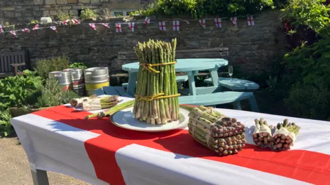 Asparagus is vertical on a plate on a table outside that is covered by a St George's flag. Other asparagus is horizontal on the table, while a light blue table, with light blue benches for seating, is in the background. 