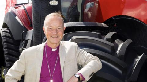 Rt Rev Dr Mike Harrison wearing Bishop's purple shirt with clerical white collar and white jacket standing in front of a tractor