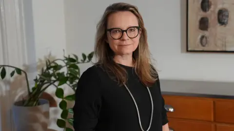 Prof Helen Duffy, international legal counsel for Abu Zubaydah, wears a black top, long white necklace, black glasses and has blonde hair. She is standing in front of a plant, wooden cabinet and painting on the wal 