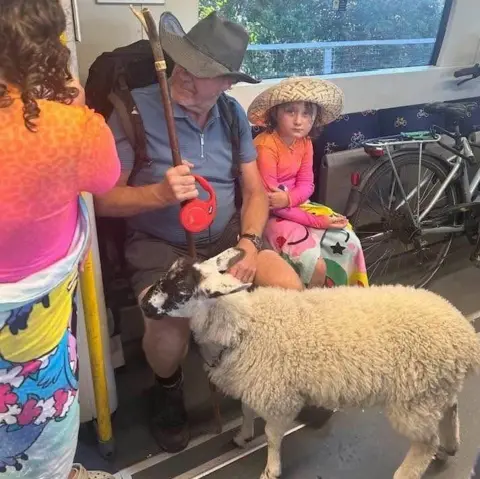 Gary Templeton Ava is sat on the train wearing a straw hat. She is wearing an orange top and colourful skirt. Jai is sat next to her with the lamb standing at his feet.