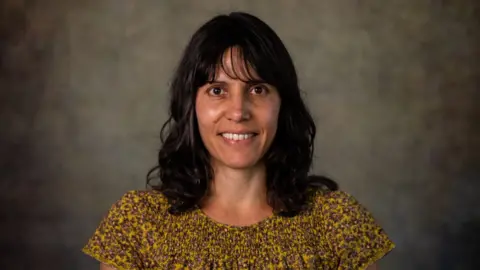 University of Reading A picture of Prof Holly Joseph, who is wearing a yellow top and has black hair. She is smiling at the camera and is standing in front of a grey background.