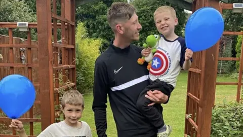 Family handout / Lincolnshire Police Laurie Ellis wearing a black sports top. He has short brown hair and is holding Mason with his left arm, who is wearing a grey Captain America T-shirt and is holding a blue balloon and dinosaur toy. Louie is holding his father's right hand and is also holding a blue balloon.