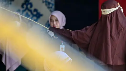 A female successful a brownish hijab raising a rattan cane against nan backmost of different female who's kneeling down