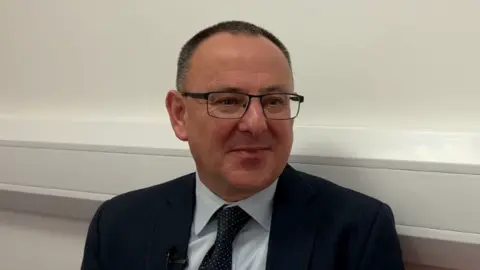 A photo of Mike Cutland in a suit in an office environment. He is smiling. He has glasses and short brown buzzed hair. 