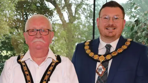 Causeway Coast and Glens Council Oliver McMullan and Richard Stewart posing with their new chains of office in front of a large window with trees in the background. McMullan has short, white hair and a white moustache. He is wearing glasses and a white, open-necked shirt. Stewart has short dark red hair and ginger stubble. He is wearing a navy suit, white shirt and a dark green tartan tie.