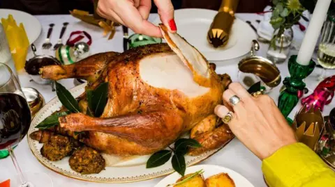 Jonathan Knowles via Getty Images Hands taking slices of meat from a cooked, golden Christmas turkey. Also on the table are vegetables and other trimmings and Christmas crackers  