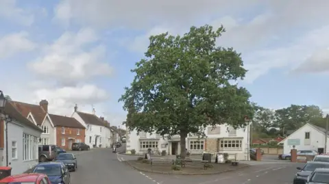 Google Maps A village square, with traditional brick houses and pub. A large green tree in the middle with benches