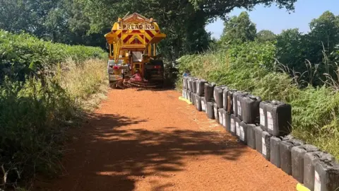 Coventry City Council A council grit lorry spreading what appears to be sand on a narrow country lane. There is a pile of oil drums piled up on the right hand side of the road. There are trees and hedgerows either side.