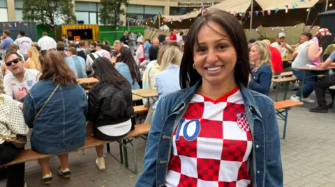 Mark Denten/BBC A smiling Croatian fan, wearing her red-and-white chequered kit shirt with a denim jacket over the top.