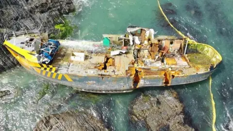A shipwreck is seen from a bird's eye view. It sits on rocks and is tilted at a slight angle. The ship has two gashes in its side.