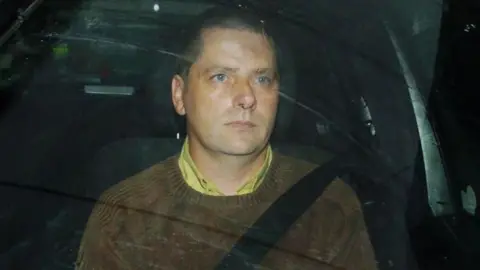 Martin McCauley is sitting in the passenger seat of a car with his seatbelt on. He has short brown hair and is wearing a brown jumper with a yellow shirt.