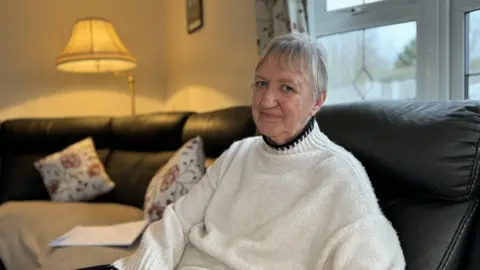 An older woman wearing a white jumper is sitting on a sofa