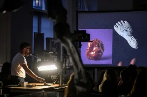Stuart Whipps Artist Stuart Whipps stands at a table in a room next to a slide projector, showing images of rocks. An audience can be seen in the darkened room. 