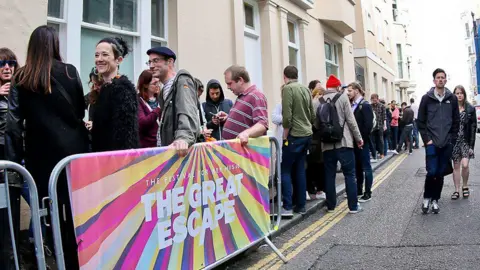 People queueing outside a row of cream houses with white windows. They are queueing behind metal fencing with rainbow netting on which reads The Great Escape. To the right are people walking towards the camera along a road