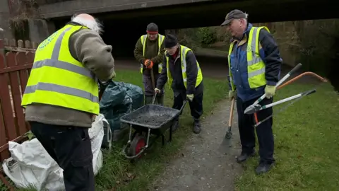 Four older men wearing yellow hi-vis jackets standing on a canal towpath. One man is holding the handles of a wheelbarrow which is filled with rubble. Another man is holding shears and another has his back to the camera. The man in the far right is looking down into the wheelbarrow and holding a tool that reaches the floor. 