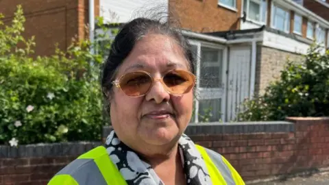 Woman with black hair wearing glasses and a high-visibility vest standing outdoors in front of a residential building with brick walls, windows, and greenery.