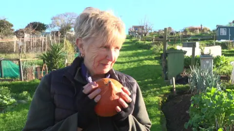 Angela is pictured holding a round, orange squash and smiling. She is wearing black, fingerless gloves, a black, padded gilet and a muted green zip-up top. Angela is standing on some grass near a wheelbarrow full of squashes, at the allotment.
