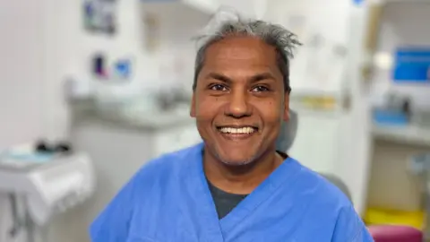 Martin Giles/BBC Lenny Lalsie is smiling and has greying black hair. He is wearing a blue scrubs top and is sitting in a dental clinic.