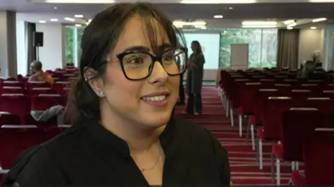 Amarjit standing in the foreground of a large conference or meeting room. She is wearing a black top, dark-framed glasses, small stud earrings, and a thin necklace. Her hair is tied back, with a few strands falling across the forehead. The room contains many rows of red upholstered chairs arranged in neat lines facing the front. A red patterned carpet with horizontal stripes runs through the room. The lighting is bright, with multiple ceiling lights illuminating the space.