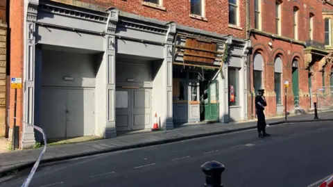 A police officer is standing on a road within a cordon outside a two-storey building. Police tape can be seen at one end of the cordon. Wooden doors to the building are open.