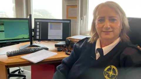 Debbie is wearing her navy blue uniform and is sitting at a chair in front of radios and computers at the watch station. She has shoulder-length blonde hair and is smiling at the camera.