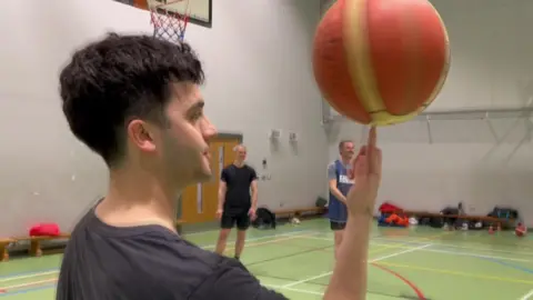 A man with short black hair and a black t-shirt spins a basketball on his finger.