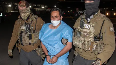 A man in a blue prison jumpsuit, Mohammad Sharifullah, wears a white surgical mask and is bound in handcuffs. Two men with obscured faces in FBI tactical gear hold his arms. It is night and there is an airplane in the background