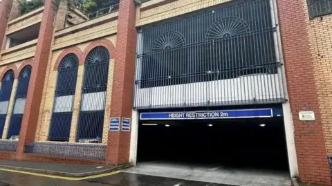 An entrance to Shaw's Brow Car park, which is brick-built with decorative iron work between archways on the outside, and a blue sign over the entrance that says height restriction 2m.