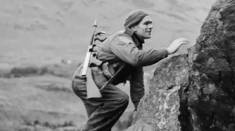 In a black and white photograph, a commando wearing combat fatigues climbs a rockface with a machine gun slung over one shoulder. There is a hill in the background.