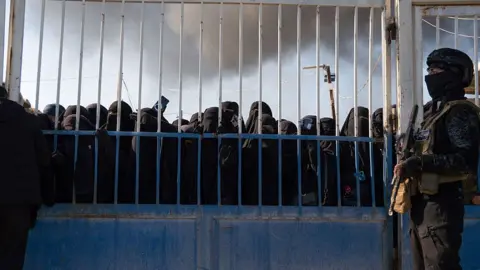 Syrian security force member pictured holding a gun and standing next to a blue metal gate at al hol camp with women in niqabs crowding at the gate
