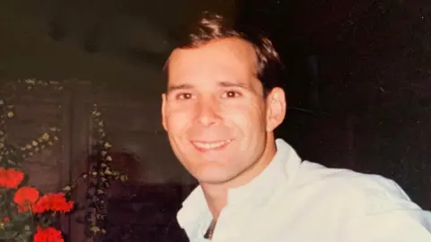 A photograph of a man stood in a garden next to red flowers wearing a white shirt. He is clean shaven, with short dark hair. 