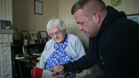 An elderly woman with white hair, glasses and wearing a blue flowered dress and grey cardigan sits in am armchair looking at a leaflet which is being shown to her by James Hunt, with sandy hair and wearing a blue hoody, who is leaning down next to her.