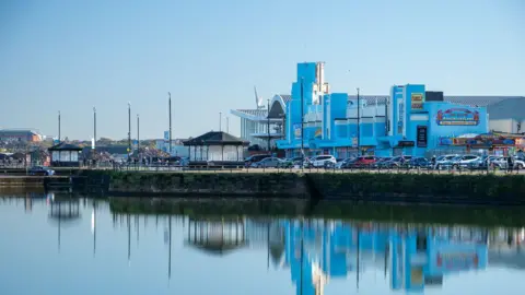 Oliver Mills/geograph Blue leisure venues can bee seen next to the car park on the promenade. There are kiosks above the water reflecting the buildings.