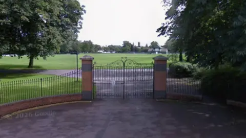 Google The entrance to a park. There is a double gate with decorative metalwork at the top. The gate is fixed to two pillars made of brick. There is a triangular piece on top of each pillar. The park comprises a large open space which is predominantly lawn. There are trees surrounding it and a cricket pavilion in the distance.
