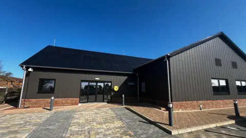 Jack Maclean/BBC The exterior of a black-clad and red brick community centre is shown under clear blue skies, with a paved area in the foreground with black bollard lights.