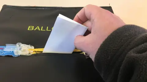 A hand is holding a white piece of paper and posting it through a ballot box. The ballot box is black with yellow writing. A large zip seals the container. 